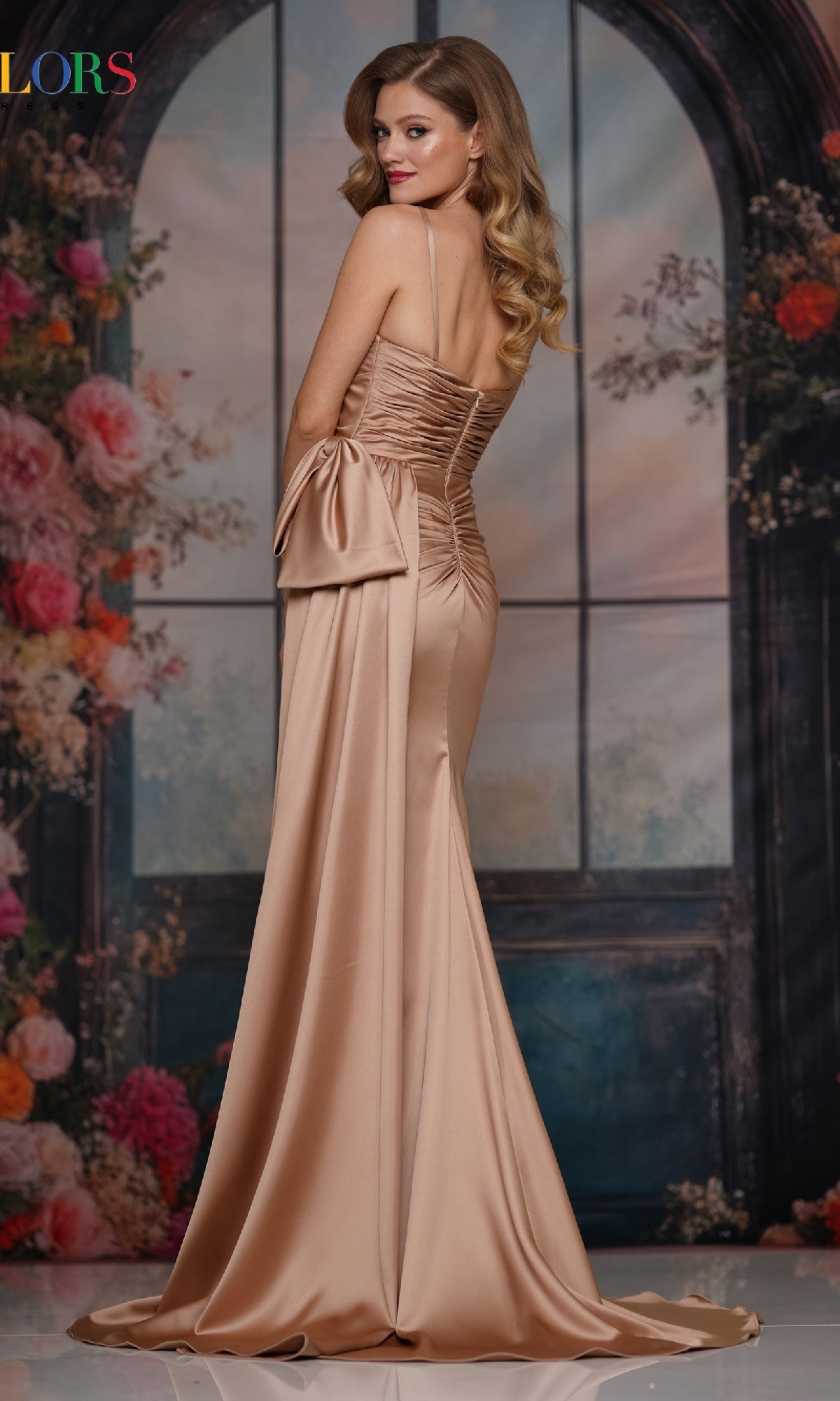 Wearing the Bow-Waist Long Satin Prom Dress: Colors Dress 3709, a woman poses before a floral arched window backdrop, showcasing its dramatic bow and train—an elegant pick for formal occasions.